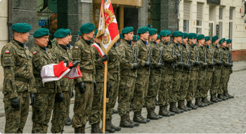 Historia AK. Zdjęcie grupy żołnierzy: Grupa żołnierzy w mundurach polowych, ustawionych w szeregu. Żołnierze noszą zielone berety, a jeden z nich trzyma sztandar wojskowy i flagę. Tło to miejskie budynki.